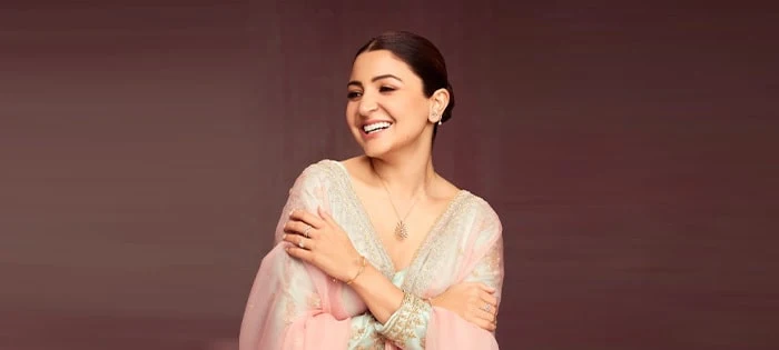 Anushka Sharma smiling and wearing a delicate GIVA pendant and stud earrings against a warm burgundy backdrop