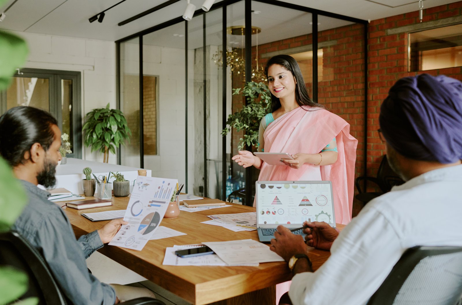 A founder in a pink saree presenting product data on a laptop to two colleagues across a wooden meeting table