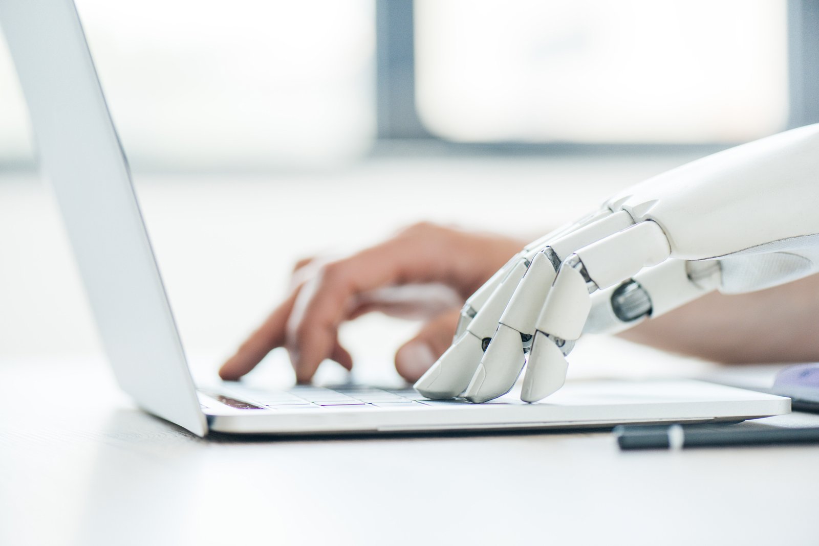 Two hands sharing a laptop keyboard — one human, one robotic — a working image of the human-plus-AI collaboration inside a modern edit suite