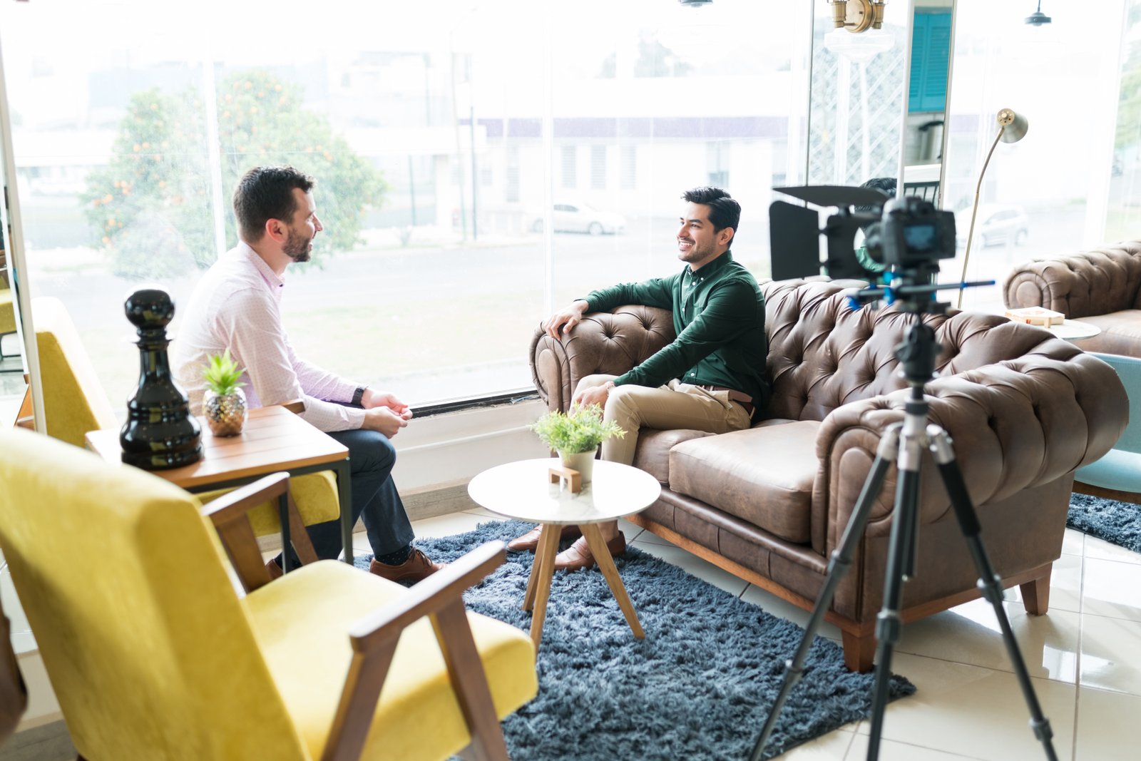 A founder mid-interview on a leather sofa, with an interviewer opposite and a camera on a tripod in the foreground