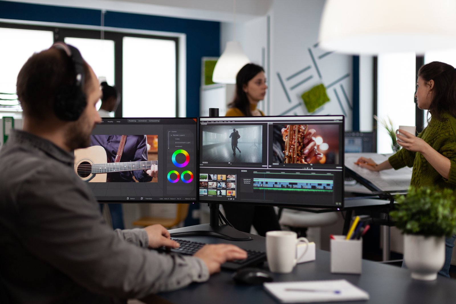 An editor at a dual-monitor edit suite — headphones on, a music track and a colour reference open — with team members reviewing the cut in the background